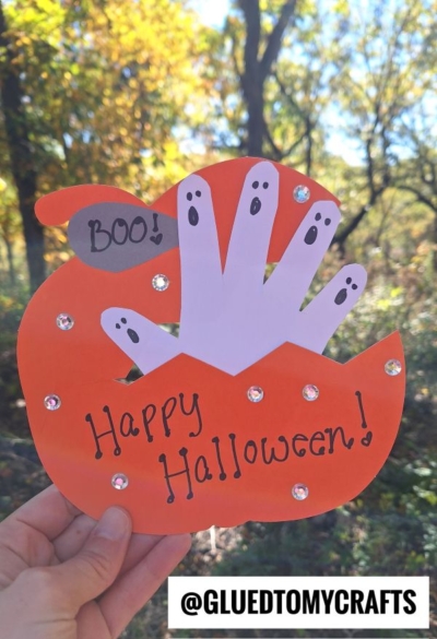 An orange paper pumpkin with a white handprint, each finger a ghost, makes the perfect halloween keepsake. “Happy Halloween!” is written on the pumpkin, while “Boo!” appears in a speech bubble. Gems decorate the festive pumpkin, with trees and sunlight in the background.
