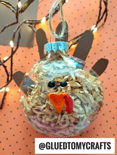 A clear turkey ornament filled with shredded paper is decorated to look like a turkey, with googly eyes, an orange beak, red wattle, and brown paper feathers. A tag at the bottom reads @GLUEDTOMYCRAFTS.