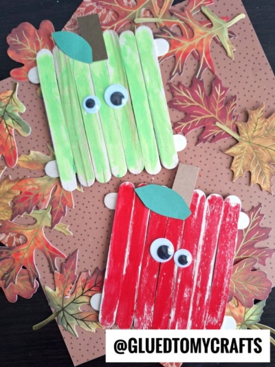 Two popsicle stick apple friends, one green and one red, with googly eyes and paper leaves, are placed on a brown, dotted autumn-themed background with paper fall leaves. @GLUEDTOMYCRAFTS is printed at the bottom.