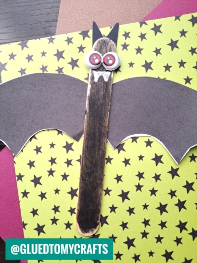 A popsicle stick bat made with a black popsicle stick body, paper wings, googly eyes, and white fangs sits on star-patterned paper. The handle @GLUEDTOMYCRAFTS is overlaid at the bottom left.