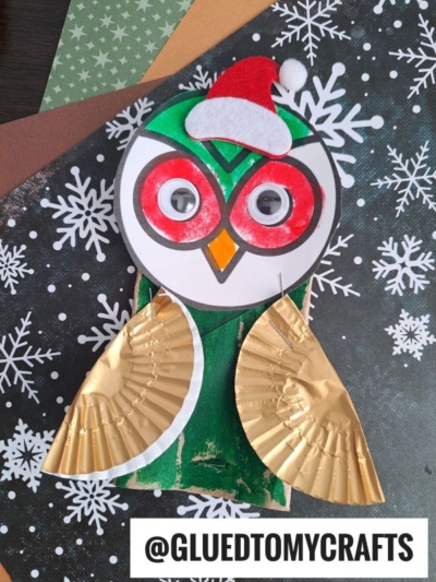 A festive paper roll owl craft features a paper face with googly eyes, a red Santa hat, and cupcake liner wings, set on a wintry, snowflake-patterned background. @GLUEDTOMYCRAFTS is written at the bottom.