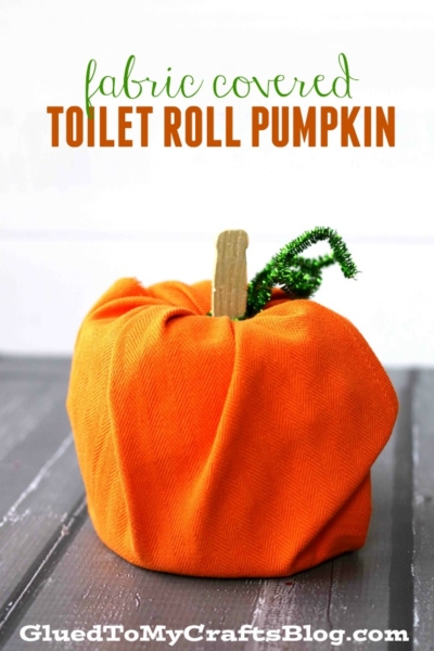 A pumpkin-shaped craft made from an orange fabric-wrapped toilet paper roll, topped with a stick for a stem and green pipe cleaner vine—an easy, fun no carve pumpkin idea. Text: “fabric covered TOILET ROLL PUMPKIN.”.