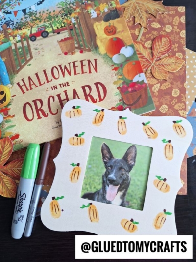 A Halloween craft setup with a thumbprint pumpkin frame holding a German Shepherd photo, autumn-themed papers, a green Sharpie, and a black pen beside a Halloween in the Orchard book.