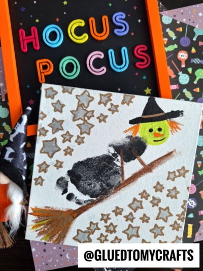 A child’s painting of a witch riding a broomstick with a green face and orange hair, surrounded by gold stars. A Hocus Pocus sign is in the background, along with decorative paper—perfect for creating treasured Halloween keepsakes.