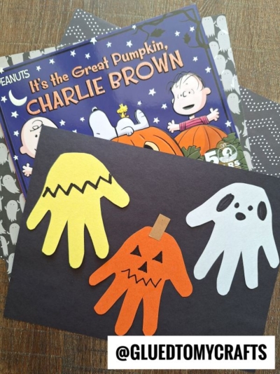 Three handprint Halloween crafts—a yellow one with zigzag lines, an orange jack-o-lantern, and a white ghost—are displayed on black paper as charming Halloween keepsakes. The book Its the Great Pumpkin, Charlie Brown is in the background.