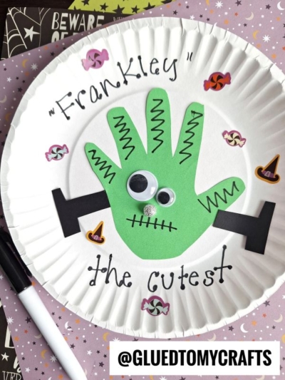 A paper plate craft of a green hand cutout as Frankley, decorated with googly eyes, black bolts, and Halloween stickers, with the phrase “Frankley the cutest” written on the plate—perfect for creating adorable Halloween keepsakes.