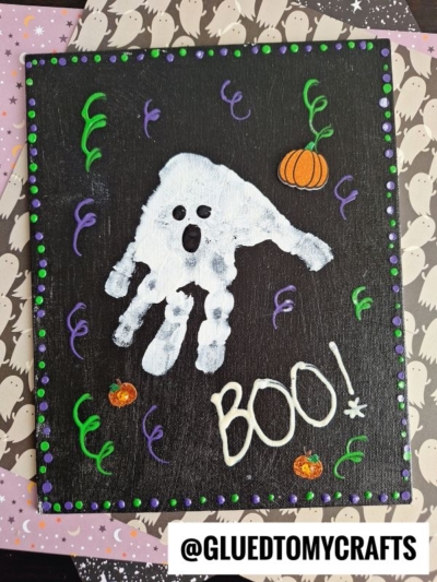 A Halloween craft with a white handprint ghost, green and purple swirls, orange pumpkins, and BOO! written in white on a black background. Perfect for creating memorable halloween keepsakes, this project sits atop ghost-patterned paper. @GLUEDTOMYCRAFTS is at the bottom.