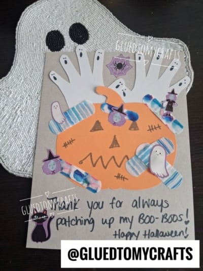 A Halloween craft with a pumpkin face, white handprints as ghosts, and bandage stickers—perfect for creating halloween keepsakes. Text reads: Thank you for always patching up my boo-boos! Happy Halloween! Instagram handle @GLUEDTOMYCRAFTS.