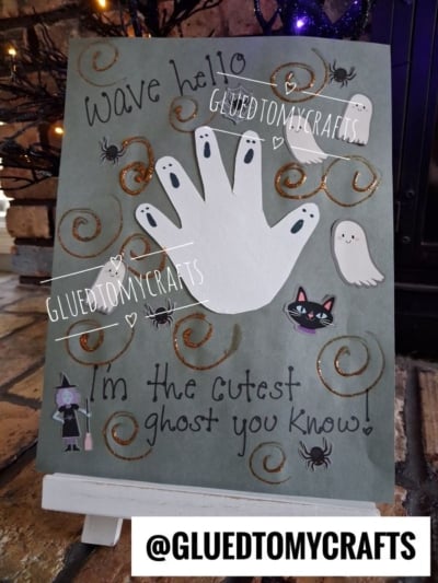 A handmade card features a white handprint ghost, cute ghost stickers, a black cat, and spiders on gray paper with “Wave hello” and “I’m the cutest ghost you know!” written in decorative text—perfect for creating cherished Halloween keepsakes.