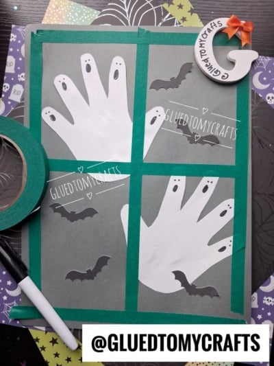 Four white paper handprints on gray paper look like ghosts, each decorated with black eyes and mouths. Black bat cutouts and a marker are nearby. Halloween-themed paper and green tape frame the crafts, making these charming creations perfect halloween keepsakes.