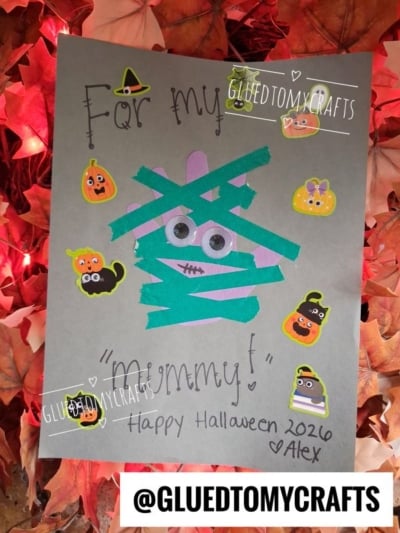 Gray Halloween card with a green mummy made of tape and googly eyes, surrounded by cute Halloween-themed stickers—perfect for halloween keepsakes. Text: For my mommy! Happy Halloween 2026. Alex. Leaf background.