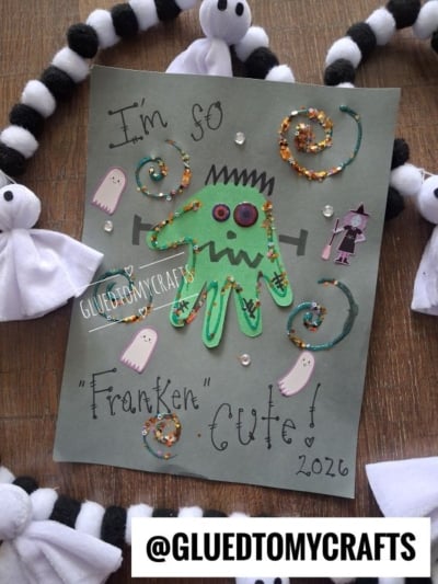 Handprint Frankenstein craft on gray paper with glitter, ghosts, pearls, and “I’m so Franken cute! 2026” written in marker. This adorable Halloween keepsake is surrounded by black and white pompom garland and tissue ghosts.