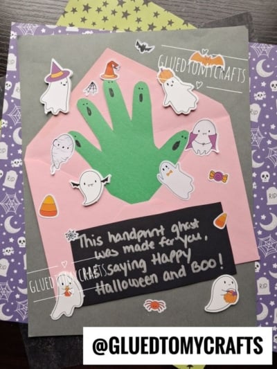 A Halloween craft featuring a green handprint cutout surrounded by ghost and candy stickers, perfect for halloween keepsakes, with a note saying, This handprint ghost was made for you—Happy Halloween and BOO!.