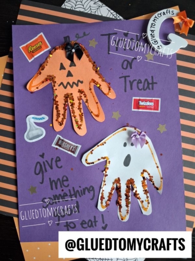A Halloween craft with orange and white handprint cutouts decorated as a pumpkin and ghost, stickers of candy, glitter, and playful handwritten messages on a purple background—perfect for creating memorable Halloween keepsakes.