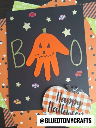 A Halloween card with BOO written in gold, an orange handprint jack-o-lantern, gold stars, candy stickers, and a plaid pumpkin cutout with Happy Halloween on orange and green patterned paper—perfect for creating memorable halloween keepsakes.