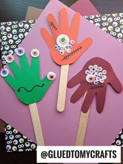 Three colorful handprint monster puppets with googly eyes and drawn mouths, made from paper and craft sticks, lay on top of patterned and solid-colored paper backgrounds—perfect as fun Halloween keepsakes. A label reads @GLUEDTOMYCRAFTS.