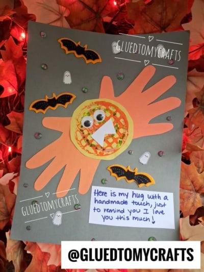 A Halloween craft features orange paper handprints, a googly-eyed circle, bats, ghosts, glitter dots, and a sweet note—perfect halloween keepsakes to remind you of hugs and handmade love!.