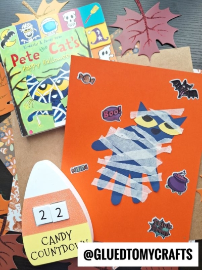 A Halloween craft with an orange card showcasing a Pete the Cat mummy made from blue paper and white tape, stickers, a Candy Countdown craft, the Pete the Cat book, paper leaves in the background—perfect for creating fun halloween keepsakes.
