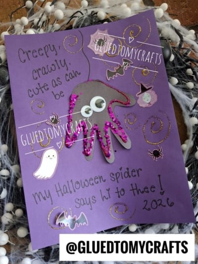 A purple card decorated with a googly-eyed spider made from a handprint, pink glitter, ghost and witch stickers, and the handwritten message: Creepy, crawly, cute as can be, my Halloween spider says hi to thee! 2026. Perfect for those who love creating memorable Halloween keepsakes.