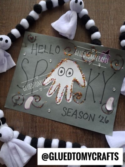 Handmade card says Hello Spooky Season with a ghost drawn from a handprint, surrounded by sparkles and ghost decorations—perfect for creating halloween keepsakes. Text @GLUETOMYCRAFTS appears at the bottom.