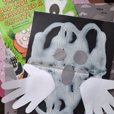 A children’s Halloween craft with paper cutouts forms a ghost with white handprint wings, placed on black paper—perfect for halloween keepsakes. Behind it are Halloween-themed books and orange bat-patterned paper. Text reads @GLUEDTOMYCRAFTS.