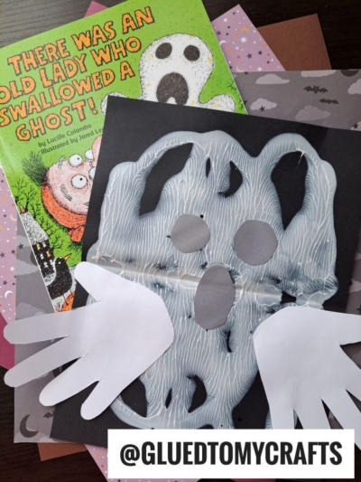 A children’s Halloween craft with paper cutouts forms a ghost with white handprint wings, placed on black paper—perfect for halloween keepsakes. Behind it are Halloween-themed books and orange bat-patterned paper. Text reads @GLUEDTOMYCRAFTS.