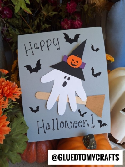 A Halloween craft featuring a ghost made from a white handprint with a witch hat, broom, and black paper bats—perfect for creating halloween keepsakes—surrounded by orange flowers and pumpkins. Text reads Happy Halloween! and @GLUEDTOMYCRAFTS.