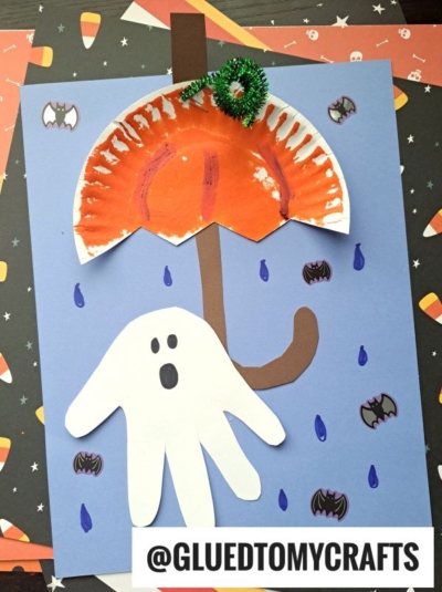 A Halloween-themed kids craft features a paper plate orange umbrella, a white handprint ghost, raindrops, bats, and a brown umbrella handle on a blue paper background—making it perfect for creating fun halloween keepsakes. GLUEDTOMYCRAFTS is printed at the bottom.