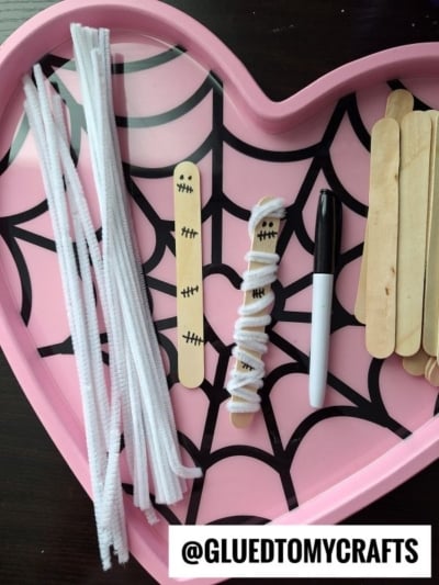 A pink heart tray with a spiderweb pattern holds white pipe cleaners, craft sticks with drawn mummy faces, a black marker, and more plain craft sticks—perfect supplies for making a pipe cleaner wrapped mummy. @GLUEDTOMYCRAFTS text is at the bottom.