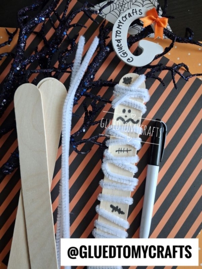 Craft supplies for a Halloween project: popsicle sticks, white pipe cleaners, a black pen, and a pipe cleaner wrapped mummy displayed on an orange and black striped background. @GLUEDTOMYCRAFTS text is shown.