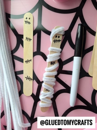 Three craft sticks on a spiderweb-patterned surface; two have black marker faces and stitch marks, while one is a pipe cleaner wrapped mummy. A white and black marker and extra white pipe cleaners are nearby. @GLUEDTOMYCRAFTS is written at the bottom.