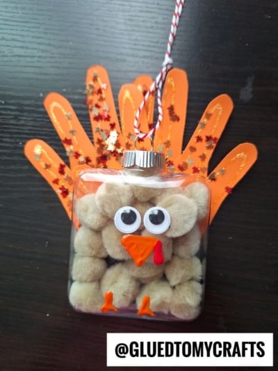 A turkey ornament craft made from a clear ornament filled with tan pom-poms, googly eyes, an orange beak, red wattle, and orange paper handprint feathers, displayed on a dark table. @GLUEDTOMYCRAFTS text is at the bottom.