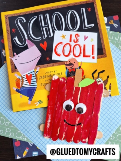 A children’s book titled School is Cool! sits beside a cheerful red popsicle stick apple friend, complete with googly eyes and a smile, displayed on patterned paper backgrounds. A tag reads @GLUEDTOMYCRAFTS.