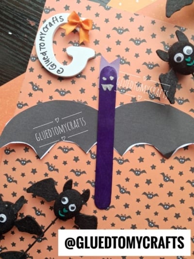 Handmade Halloween bat crafts on an orange bat-patterned paper, featuring black bats made from felt and paper, a purple popsicle stick bat craft (also known as a craft stick bat), and @GLUETOMYCRAFTS printed on parts and in bold at the bottom.