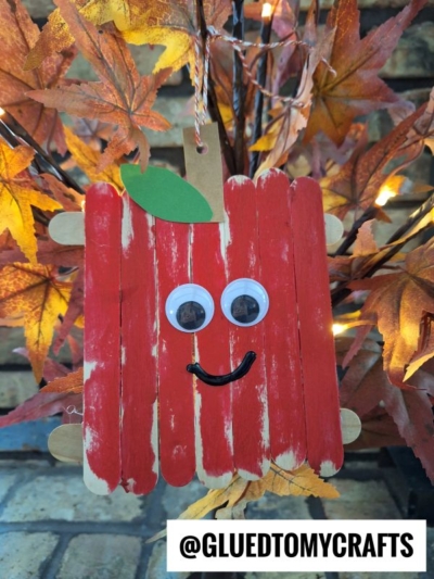 A popsicle stick apple friends decoration, with googly eyes, a green paper leaf, and a smiling face, hangs in front of autumn leaves. The handle @GLUEDTOMYCRAFTS appears at the bottom.