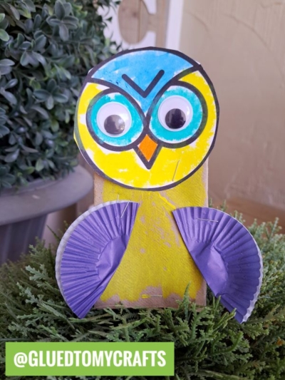 A colorful paper roll owl made from a cardboard tube, blue and yellow paper, large googly eyes, and purple cupcake liners for wings sits on green plants. A green label at the bottom reads @GLUEDTOMYCRAFTS.
