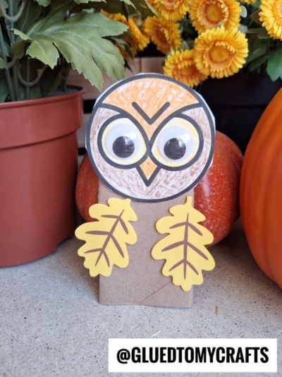 A handmade paper roll owl craft made from brown paper, with large googly eyes, drawn features, and yellow paper leaves for wings, sits among orange pumpkins and yellow flowers. Text reads @GLUEDTOMYCRAFTS.