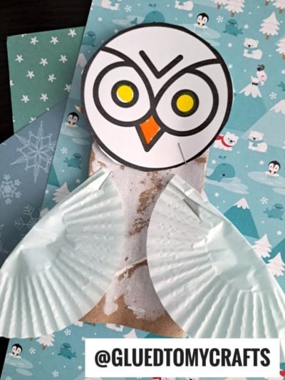 A handmade paper roll owl craft with a round paper head, yellow eyes, orange beak, and wings from white cupcake liners, set on a blue winter-themed background. @GLUEDTOMYCRAFTS is written at the bottom.