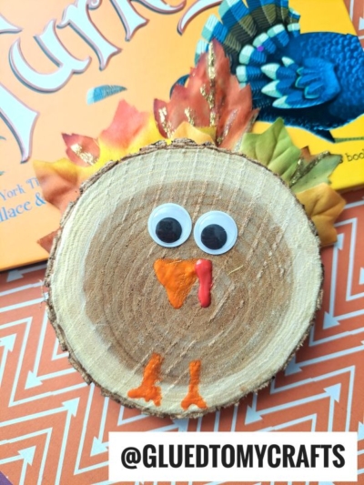 A charming wood slice turkey craft made from a wood slice with googly eyes, orange and red beak details, and fabric leaves as feathers, placed on an orange patterned surface. A turkey book is partially visible in the background.
