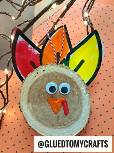A charming wood slice turkey is decorated with googly eyes, colorful paper feathers, an orange beak, and a red wattle. An orange string is attached for hanging. The background features cheerful orange dots and glowing string lights.