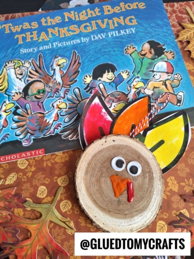 A children’s book titled “’Twas the Night Before Thanksgiving” is shown with a wood slice turkey craft, complete with googly eyes and colorful paper feathers. The handle @GLUEDTOMYCRAFTS appears at the bottom.