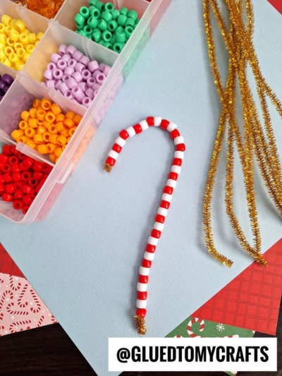 A beaded candy cane ornament made of red and white candy cane beads rests on a blue surface next to gold pipe cleaners and a box of colorful beads. The bottom right has the text @GLUEDTOMYCRAFTS in bold.