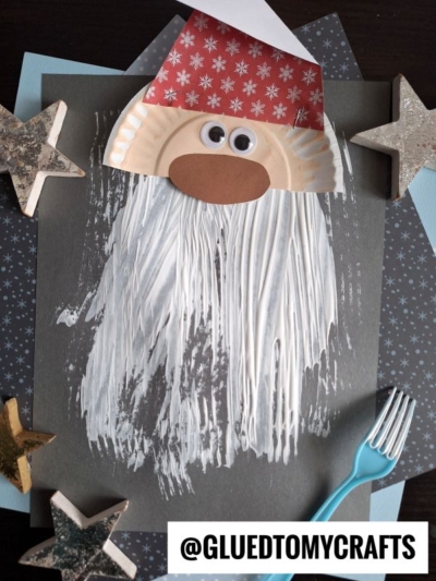 A fun fork painted Santa craft featuring a paper plate face, red paper hat, and a fork painted beard with white paint. Displayed on dark paper with star decorations. Text reads @GLUEDTOMYCRAFTS.