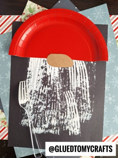 A fork painted Santa beard craft made with a red paper plate hat, brown paper nose, and white paint streaked for the beard on black paper. Find more ideas from @GLUEDTOMYCRAFTS. Perfect for your own fork painted Santa!.