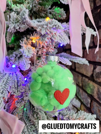 A clear grinch pom pom ornament filled with green pom-poms and decorated with a red heart hangs on a snow-flocked Christmas tree adorned with colorful lights and pink ribbon. The handle @GLUEDTOMYCRAFTS is visible at the bottom.