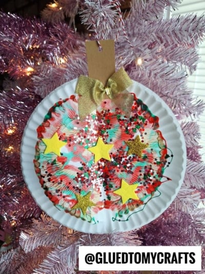 Paper plate Christmas ornament craft with colorful paint, glitter, and star stickers reminiscent of playful paint splat Christmas ornaments, topped with a gold bow and tag, hanging on a pink Christmas tree. Text @GLUEDTOMYCRAFTS displayed at the bottom.