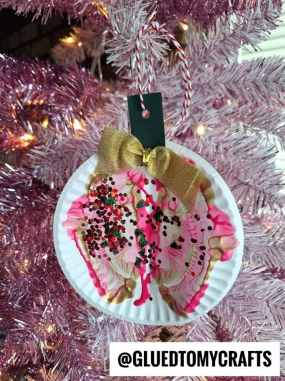 A handmade paint splat Christmas ornament made from a paper plate with pink, gold, and red paint, glitter, a gold bow, and a black tag hangs on a pink Christmas tree. @GLUEDTOMYCRAFTS is written at the bottom.