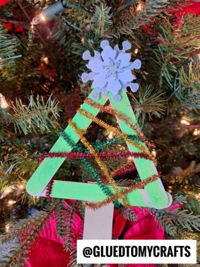 A handmade pipe cleaner Christmas tree ornament, crafted from green-painted popsicle sticks and adorned with gold and red pipe cleaners plus a purple snowflake, hangs on a tree. Text at the bottom reads @GLUEDTOMYCRAFTS.