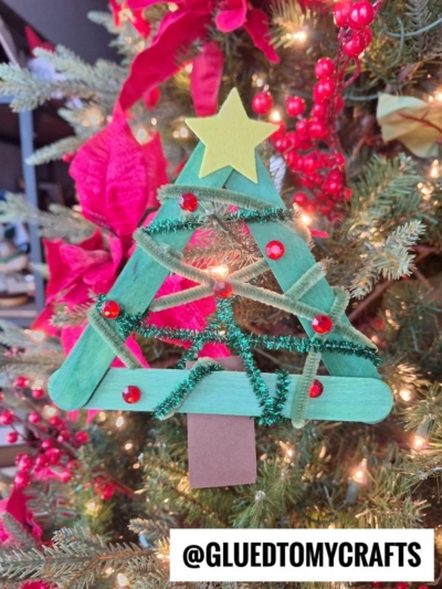 A handmade pipe cleaner Christmas tree ornament crafted from green popsicle sticks, green pipe cleaners, red pom-poms, and a yellow star, hangs on a decorated Christmas tree. Text at the bottom reads @GLUEDTOMYCRAFTS.