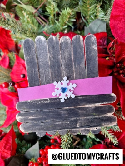 A handmade popsicle stick snowman hat, decorated with a purple paper band, a white snowflake with a blue center, and surrounded by red poinsettias and greenery. Inspired by Frosty the Snowman. @GLUEDTOMYCRAFTS is at the bottom.